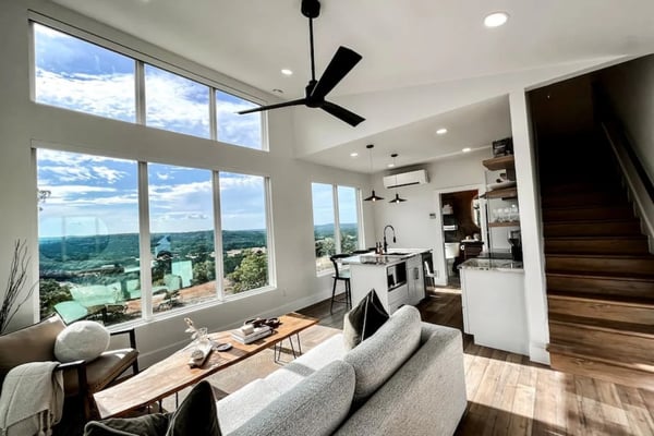 Modern open-plan living room with floor-to-ceiling windows overlooking forested valley landscape, gray sofa, kitchen island, and black ceiling fan.
