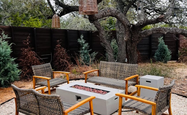 Outdoor seating area with four woven chairs around a fire pit table beneath a large gnarled oak tree with dark fence behind