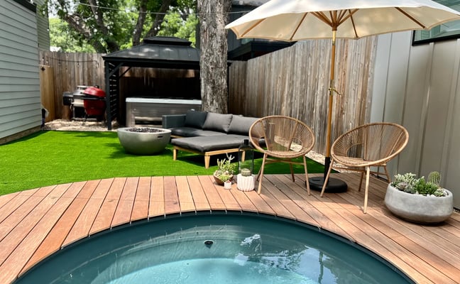 Backyard with round soaking tub, wooden deck, woven chairs under umbrella, lounge seating, fire pit, and grill